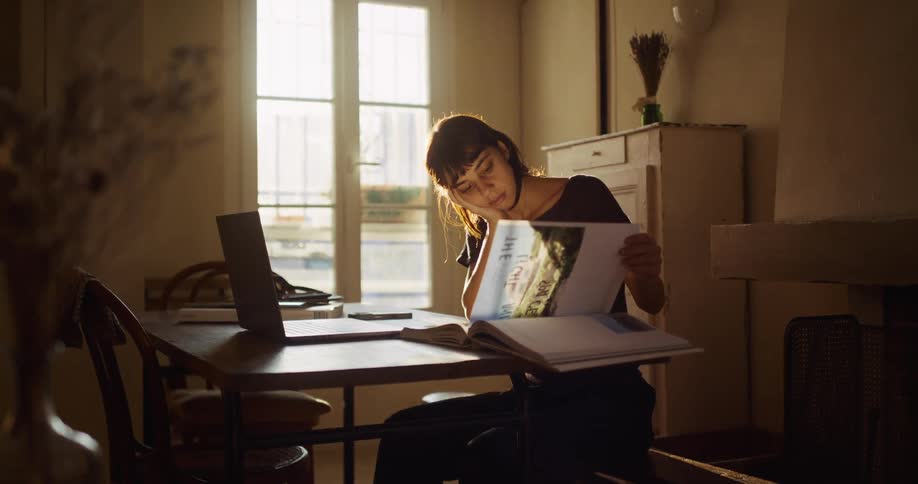 A focused young woman studies a book for inspiration before working on her laptop in her sunlit home office