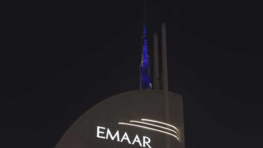 A night shot slowly reveals the iconic Burj Khalifa, illuminated in brilliant blue, from behind the glowing Emaar building in Dubai