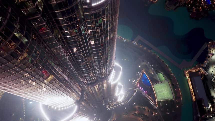 A vertiginous top-down view of an illuminated modern skyscraper at night, towering over the city lights and a vibrant urban landscape below