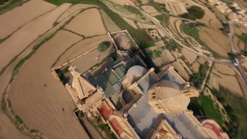 A dynamic FPV drone flight soars and dives over the magnificent Ta' Pinu Basilica in Malta, capturing its stunning architecture and the surrounding terraced fields