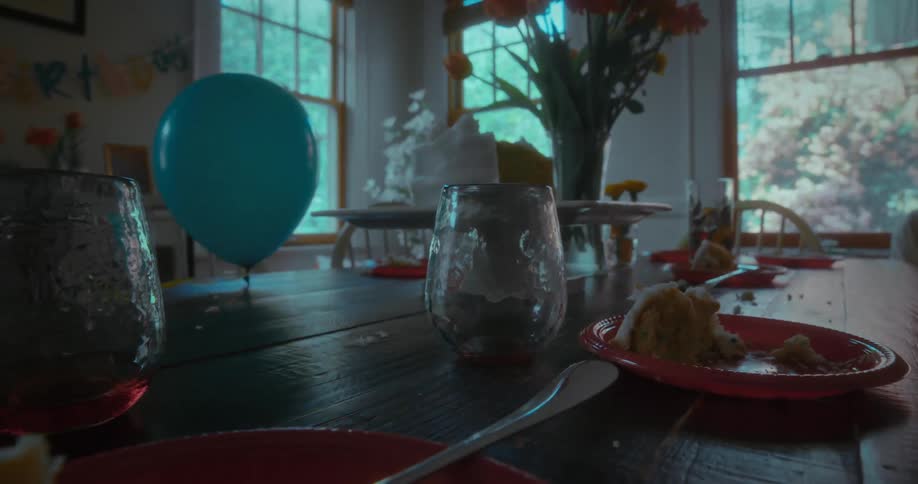 A moody, low-angle shot of the aftermath of a birthday party, featuring leftover cake and a balloon on a messy dining table