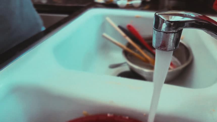 A person washes a red bowl and spatulas in a sudsy kitchen sink with a running faucet in this slow-motion, close-up shot