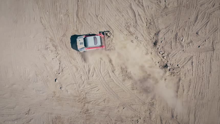 An aerial top-down slow-motion view shows a skid-steer loader backing up and moving dirt on a construction site, kicking up dust