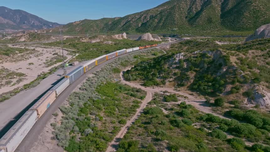 Spectacular aerial footage follows a long freight train as it navigates a winding track through a vast, sunlit desert valley