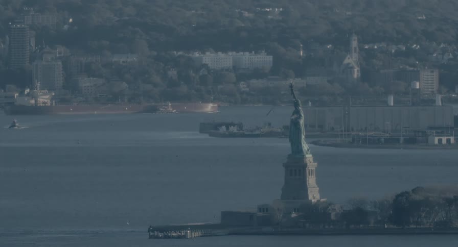 A static, telephoto shot captures the Statue of Liberty in the hazy New York Harbor as ships pass in the background