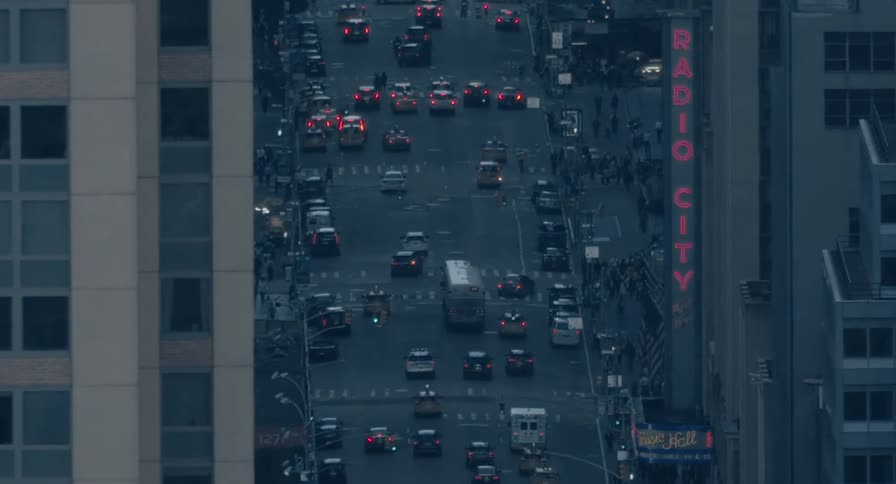 A high-angle view captures the bustling evening traffic and pedestrians on a street in New York City, with the iconic red Radio City sign glowing at dusk