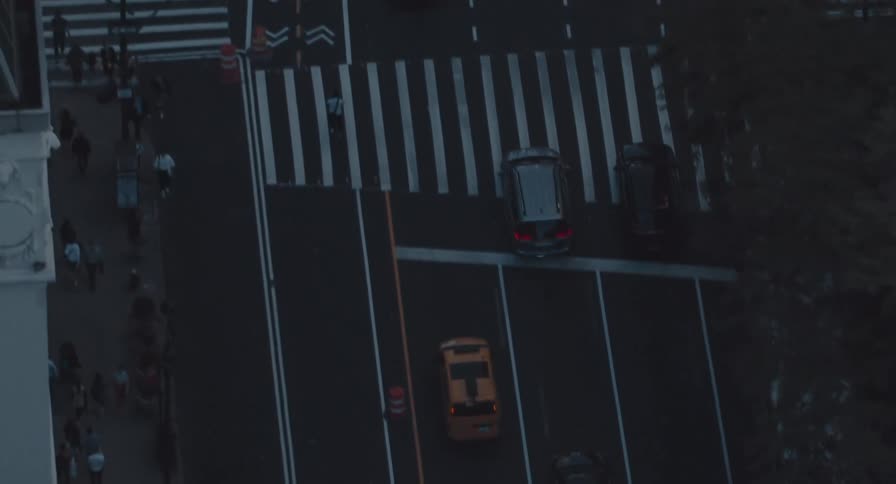 An aerial top-down view tracks yellow cabs and cars driving through a moody New York City street intersection at dusk