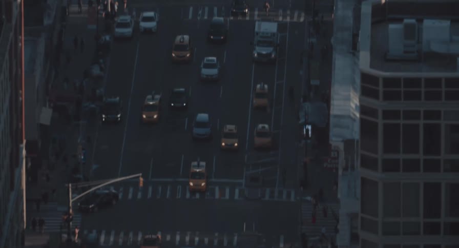 Birds-eye view of a bustling city street at dusk, where traffic, including iconic yellow cabs, moves through intersections as people walk on sidewalks