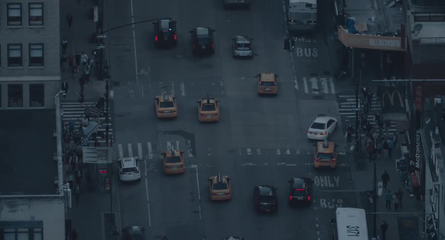 A high-angle, birds-eye view of a busy city street intersection at dusk, capturing the constant flow of traffic and pedestrians