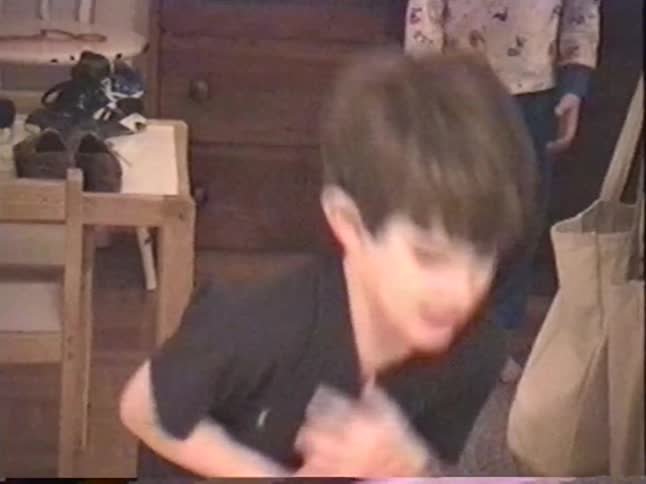 A young boy playfully tosses a yellow ball for the camera while his brother watches in the background in a nostalgic home video