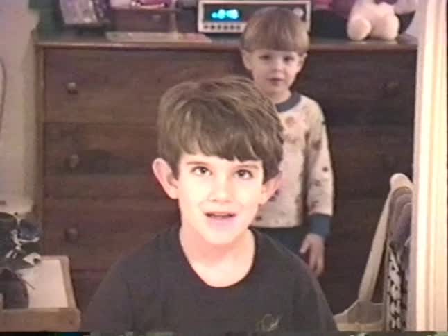A vintage home video shows a happy young boy in an insect shirt smiling at the camera while his little brother stands in the background