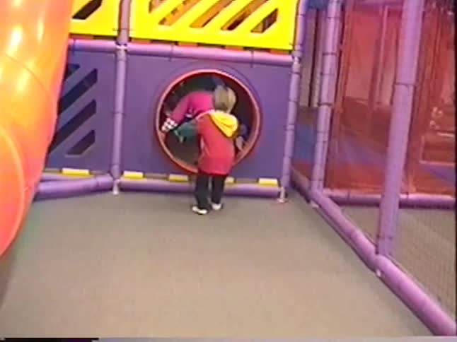 A little boy and girl happily run through a colorful indoor playground in nostalgic home video footage