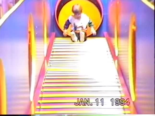 A young boy smiles as he slowly comes down a colorful roller slide at an indoor playground in a vintage home video