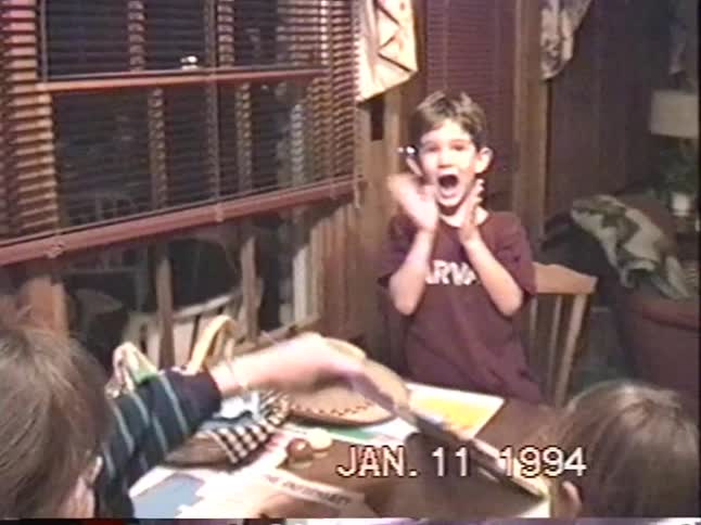 An excited young boy in a Harvard t-shirt claps and yells with joy as he opens a gift in a 1994 home video