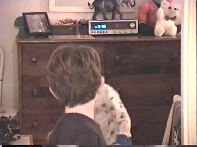 A young boy smiles happily for a vintage home video camera as his toddler sibling crawls on the floor behind him