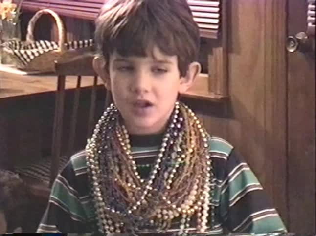 A nostalgic home video from the 90s shows a young boy smiling while wearing a huge pile of Mardi Gras beads