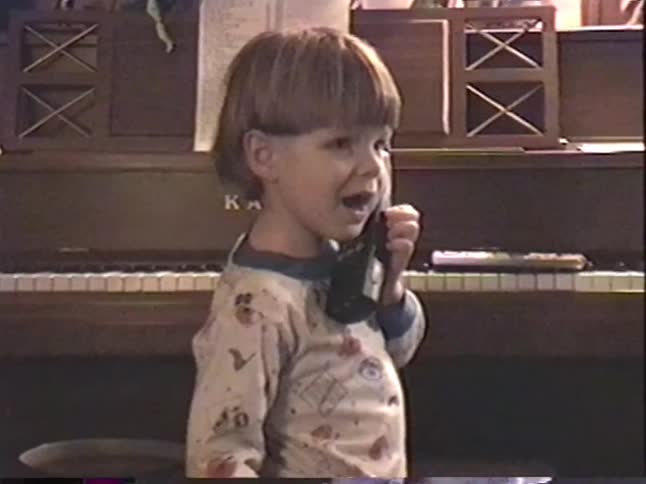 An adorable boy in his pajamas smiles while having a pretend conversation on a 90s cordless phone