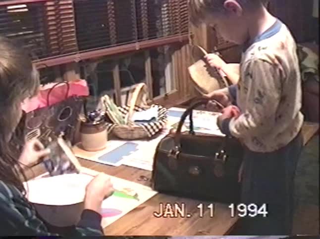 A vintage 1994 home video shows children playing together, putting toys and a cowboy hat into a handbag at a wooden table