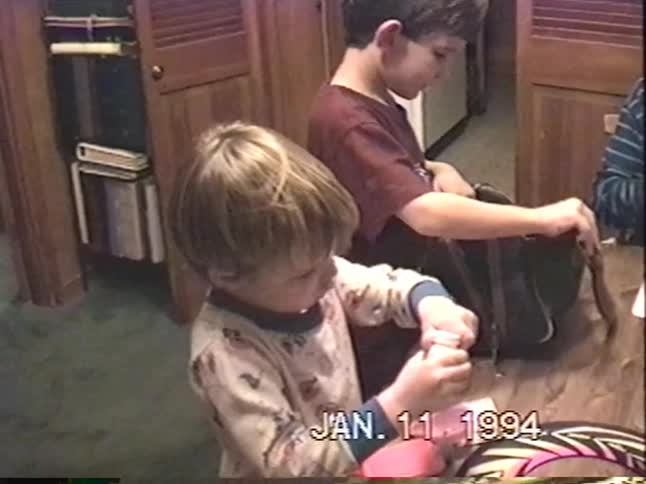 A vintage home video from 1994 shows two young brothers playing with toys at a table as their mother puts a toy hard hat on one of their heads