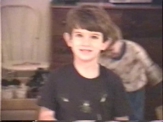 A vintage home video shows a young boy in a bug-themed t-shirt waving and making silly faces for the camera while his younger brother laughs in the background