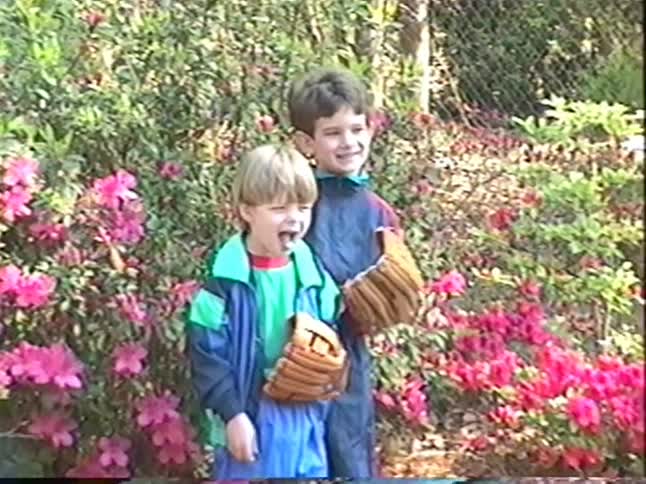 Two young brothers in colorful 90s tracksuits pose with their baseball mitts in front of a flowering azalea bush