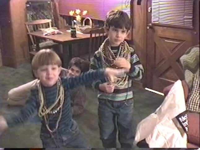 Three young boys covered in Mardi Gras beads playfully wave and dance for the camera in a nostalgic home video