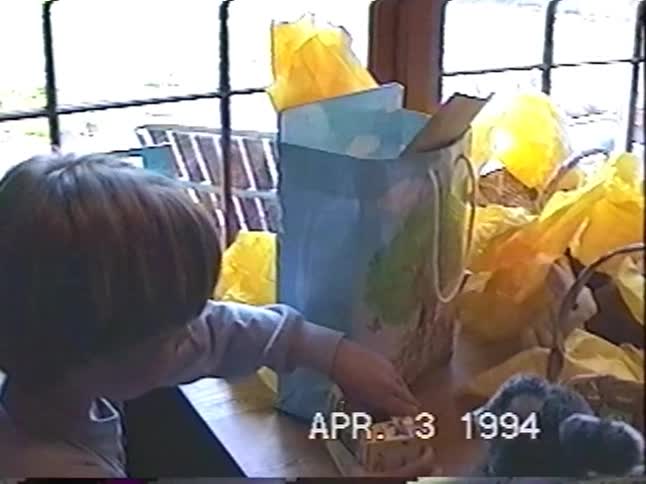 A young child explores their Easter baskets and gifts on a table in a nostalgic home video from 1994