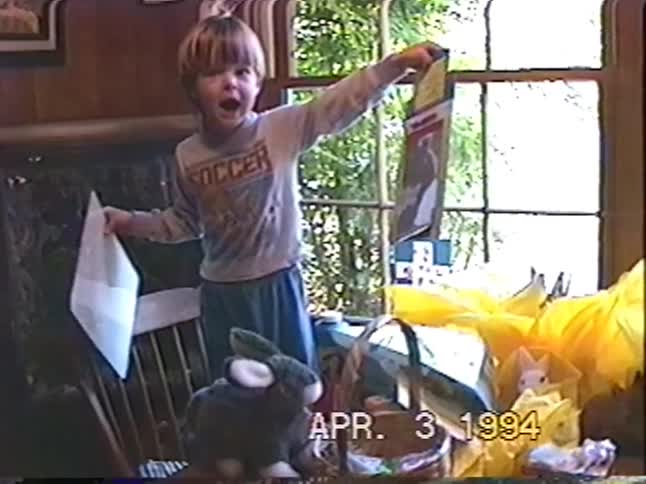 An excited young boy in his pajamas shows off his Easter gifts by the window in a nostalgic home video from 1994