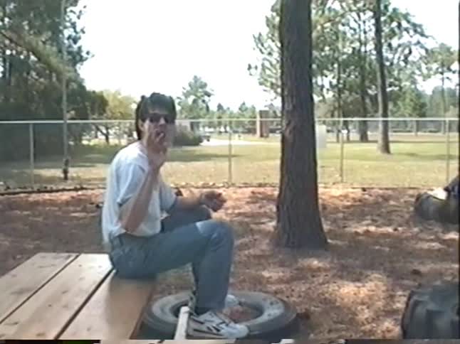 Vintage home video footage shows a man talking and eating on a bench as a young boy plays on a rope swing in a park
