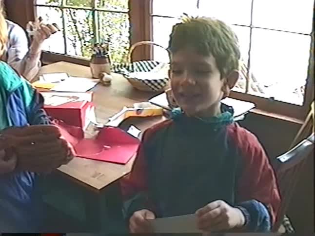 A young boy happily opens a greeting card at a table while his brother watches in a nostalgic home video from the 1990s