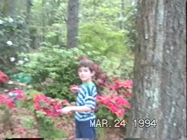 A vintage VHS recording from 1994 shows a young boy watering colorful azaleas in a beautiful spring garden