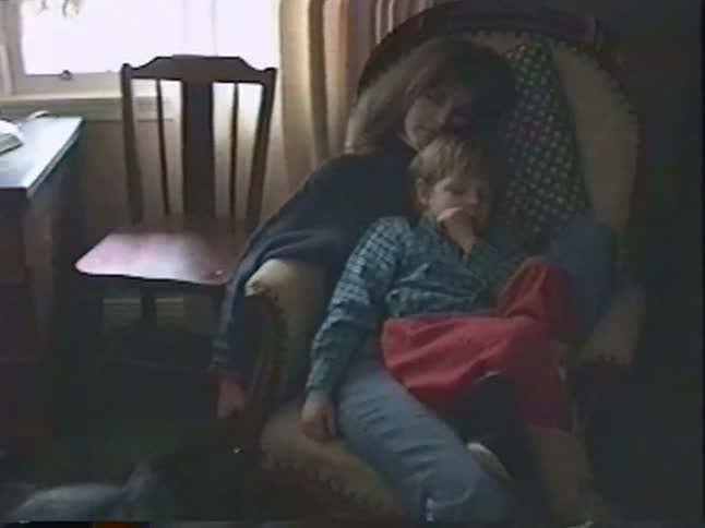A tender moment from a vintage home video shows a mother holding her sleepy son in a cozy chair as their dog rests nearby