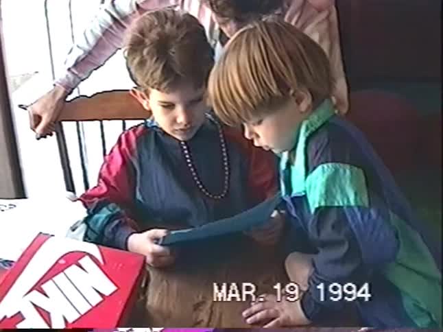 Two young brothers in colorful 90s windbreakers read a birthday card together at the dining table in this sweet home video from 1994