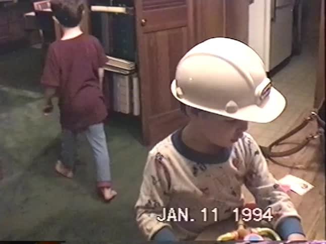 A young boy in pajamas and a construction helmet plays with a toy doctor kit at a table in a 1994 home video