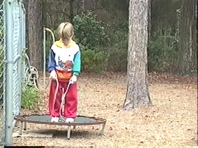 Vintage home video shows two black dogs wrestling and two young boys playing with a jump rope and a trampoline in their backyard
