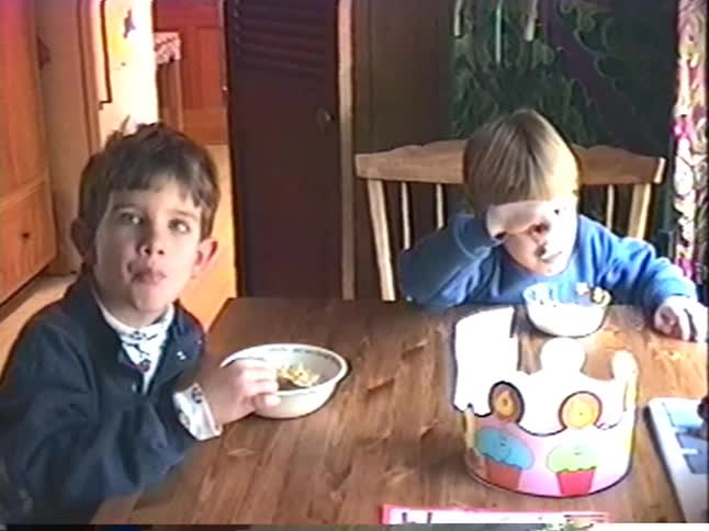 Vintage VHS footage shows two young brothers eating breakfast at a wooden table before panning to the kitchen