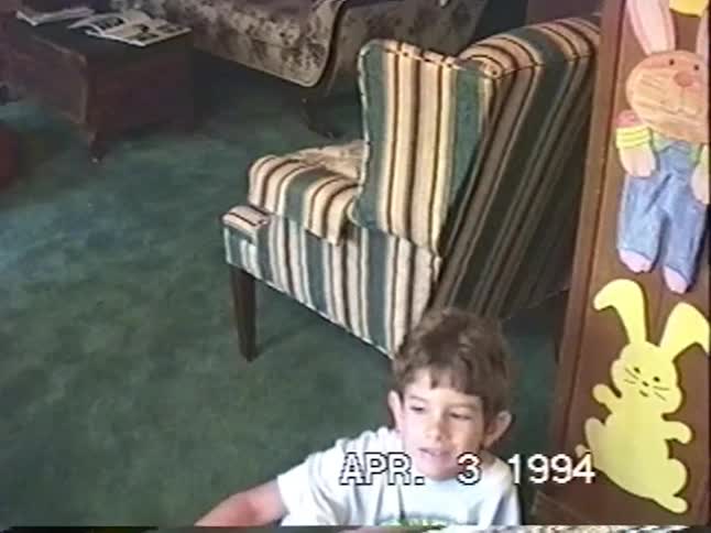 A young boy throws a colorful koosh ball at the camera in a 1990s living room with Easter decorations