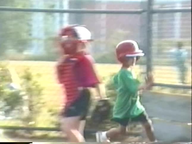 A young boy in a green shirt and red helmet runs the bases during a youth baseball game in a nostalgic home video