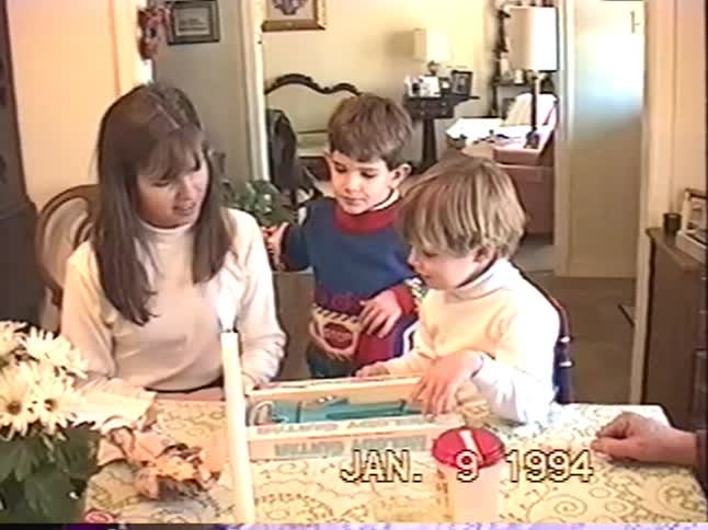 A mother helps her young sons open a toy guitar for a gift at the dining table in a nostalgic home video from 1994