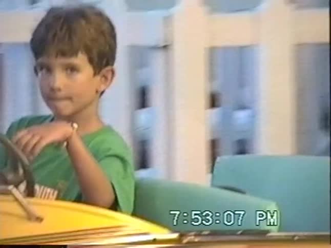 Nostalgic home video footage shows a young boy in a green shirt happily riding a spinning amusement park ride