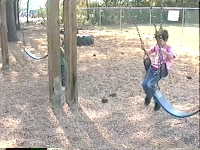 Vintage VHS footage shows a young boy having fun at a wooden playground, sliding down a pole and using the swing set on a sunny day