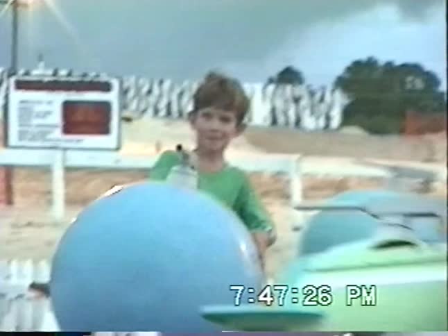 A vintage VHS home video captures children having fun on a spinning rocket ship ride at an amusement park