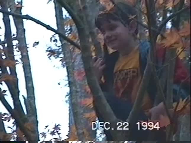 A young boy smiles and talks while perched in an autumn tree in a nostalgic home video from 1994