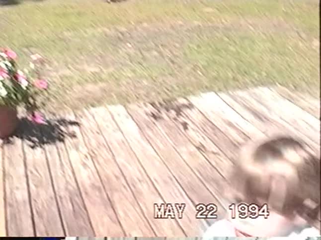 A young boy with a towel cape watches his sibling run through a sprinkler before joining in the fun in this 1994 home video