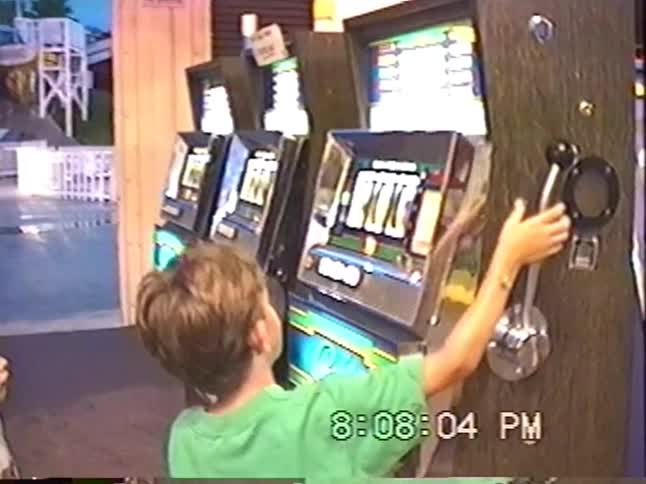 A young boy in a green shirt pulls the lever on a classic slot machine in an arcade, captured on vintage home video