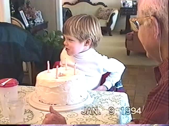 A young boy blows out the candles on his birthday cake as his family watches in this vintage home video from 1994