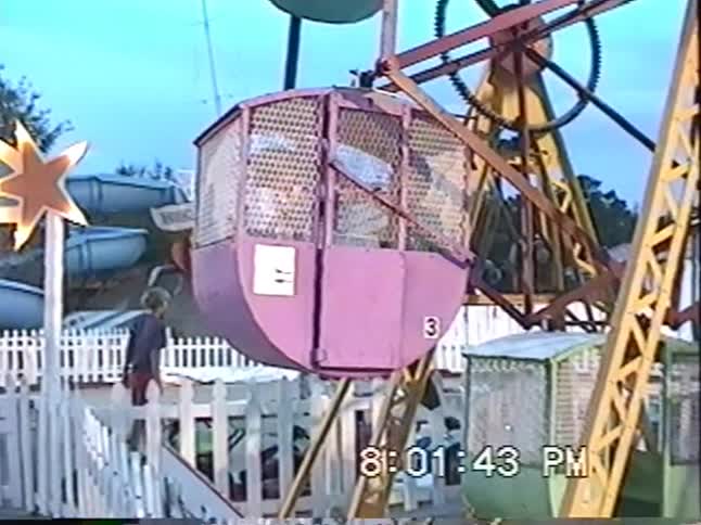 A vintage home video captures a colorful old-fashioned Ferris wheel spinning at a carnival at dusk