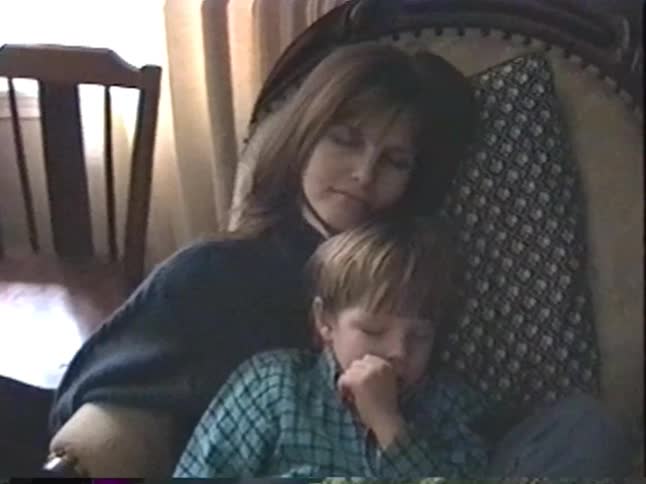 A mother lovingly holds her sleeping son as they nap together in an armchair in a cozy 1990s home