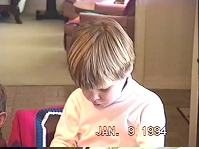 An adorable little boy unwraps a gift from his grandfather at the dining room table in a vintage 1994 home movie