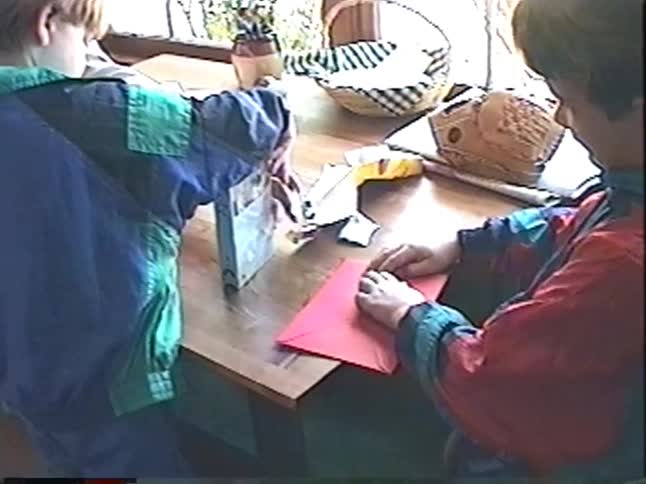 Two young boys in 90s attire unwrap gifts, one receiving a VHS movie and the other opening a card, at a wooden table in their home